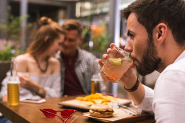 Cold drink. Serious young man drinking his cold cocktail while two of his friends flirting