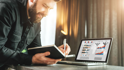 Bearded smiling businessman is standing by laptop, writing in notebook. Entrepreneur analyzes information, compares data