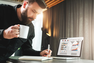 Bearded smiling businessman is standing by laptop, writing in notebook. Entrepreneur analyzes...