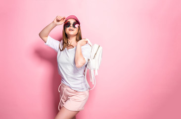 Attractive young female in stylish outfit touching cap and looking up while standing on pink background