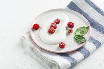 plate with homemade yogurt and ripe raspberries on a white table with green leaves