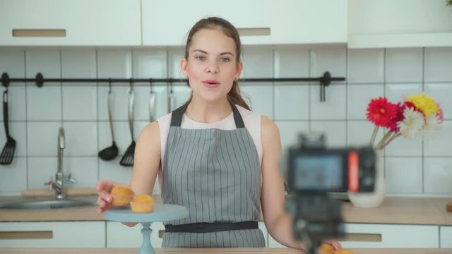 Young woman in a gray apron leads a culinary blog and records video in light kitchen. Vlogger welcomes the followers. Video shooting of the blog. Modern marketing, social networks.