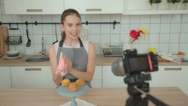 Modern woman shoots a video of the process of making dessert - a capcake confectionery vlog. Culinary blog: young woman decorates a capcake with a cream, using a pastry bag, and comments her actions.