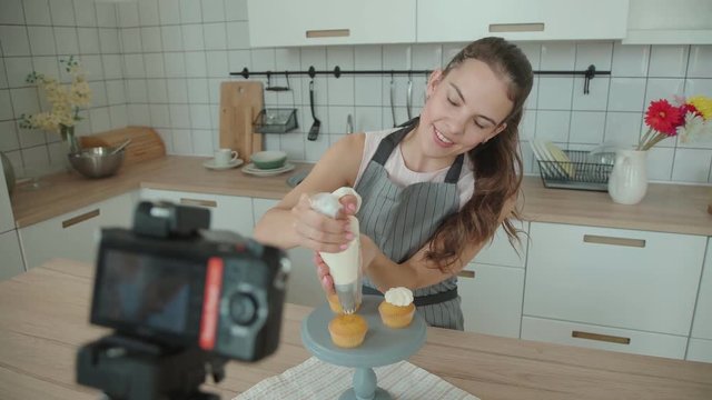 Beautiful young confectioner in apron records a video lesson, master class, video with the process of preparing a capcake and decorating cup-cake with cream. Shooting video blog or vlog