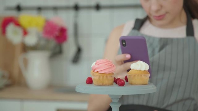 Appetizing capkakes with white and pink cream on a cake stand. The confectioner sprinkles them with decor and decorates with berries, then takes pictures on a smartphone. Culinary blog.