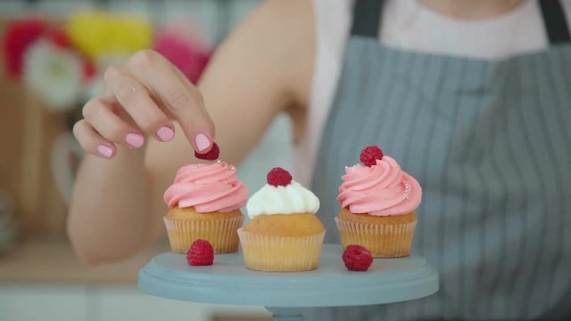 Appetizing capkakes with white and pink cream on a cake stand. The confectioner sprinkles them with decor and decorates with berries. Three cakes. In the frame, only the hands of a pastry cook