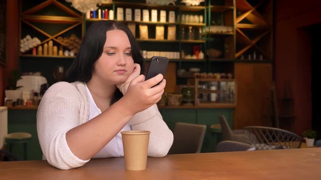 Heavy Caucasian Female Swiping Her Phone And Sitting In The Cafe With Big Paper Cup