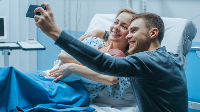 Father Takes Selfie of Him with His Wife Holding Newborn Baby while Lying on the Hospital Bed. Happy Young and Smiling Family.
