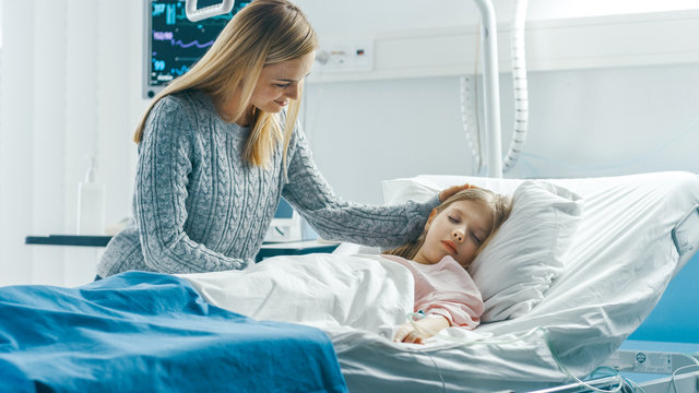 Cute Little Girl Sleeps on a Bed in the Children's Hospital, Caring Mother Covers Her with a Blanket and Caresses Her Forehead. Modern Pediatric Ward.