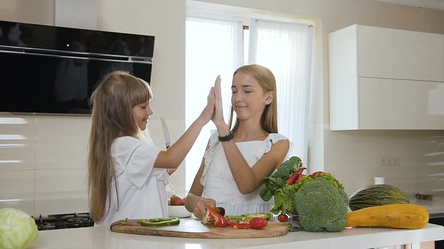 Two sisters with long hair in white dress are cutting fresh vegetables on a wooden bench in a modern kitchen. Attractive girls are cutting green and red pepper paprika and giving each other five after
