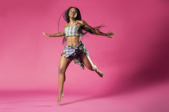 Beautiful African Black girl wearing traditional colorful African outfit does a dramatic dance move against a colorful pink background