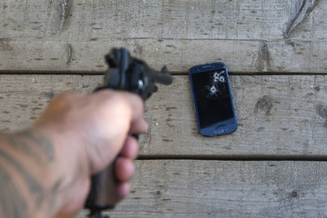 A man's hand with a pistol shoots at a mobile phone.
