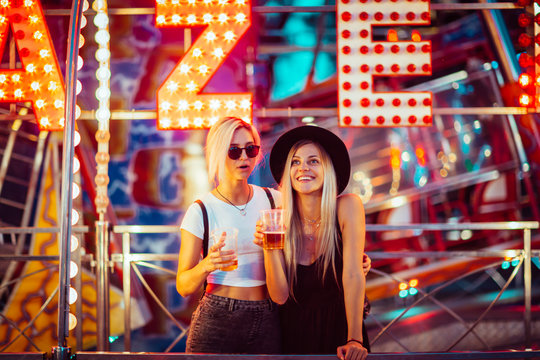 Happy female friends in amusement park drinking beer. Two young women enjoying night at amusement park. 