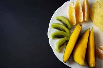 Tropical fruits, kiwi, mango, pineapple, orange and mandarin orange slices placed on white plate isolated on black background. Top view, flat lay, half plate aligned to right. Space for text.