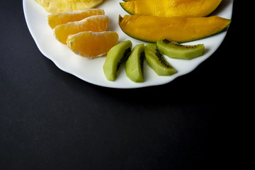 Naklejka na ściany i meble Tropical fruits, kiwi, mango and mandarin orange slices placed on white plate at the top in half, isolated on black background. Top view, flat lay.