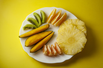 Tropical fruits, kiwi, mango, pineapple, orange and mandarin orange slices placed on white plate isolated on yellow background. Top view, flat lay.