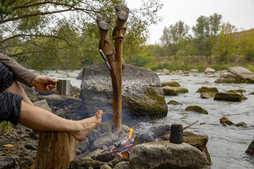A man is warming himself by the campfire.The shoes are dried. Military boots. Tourist shoes. A tourist camp on the river bank. A halt on the bank of the river. Tourist equipment.