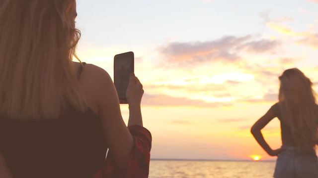 A young girl enjoys the view of the sea on the beach and takes pictures on the phone other girl. Silhouetted woman at sunset beach. Silhouettes at sunset.