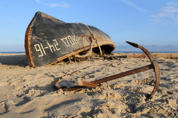 A black fishing boat lies upside down with a keel on a sandy beach. Anchor lies on the sand next to the ship.