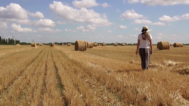 Beautiful calm woman walking around summer countryside wheat field full of bales of hay in slow motion on hot sunny day