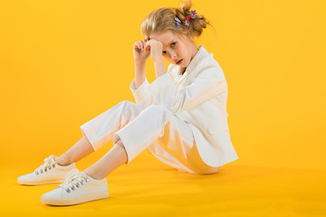 A teenage girl in white clothes sits stretching her legs forward on a yellow background.