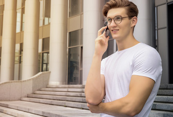 Young man student wearing glasses and a white T-shirt, sitting on the street and talking on the phone