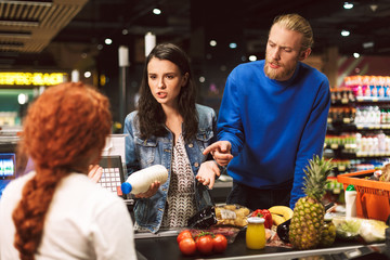 Young woman with milk of bottle in hand and man emotionally talking with cashier in supermarket
