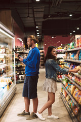 Fototapeta na wymiar Young guy and pretty girl standing back to back choosing products while spending time in modern supermarket