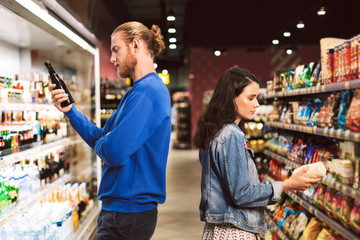 Young thoughtful couple standing back to back choosing products while spending time in modern supermarket