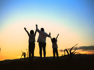 Silhouette of happy mother and two children standing with raised hands on the mountain at the sunset time.