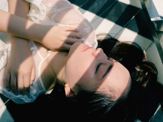 Top view of a sexy girl in white dress lying on ground with shadows on face in sunny day, arms crossed on chest and eyes closed, close up portrait of sleepy face.