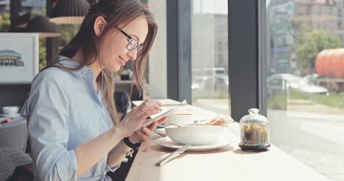 Portrait of Young Woman Using Smart Phone in a Cafe by the Window. 4K SLOW MOTION, DCi, Dolly Shot. Attractive business woman, using cellphone in the modern bistro during lunch. Technology in life