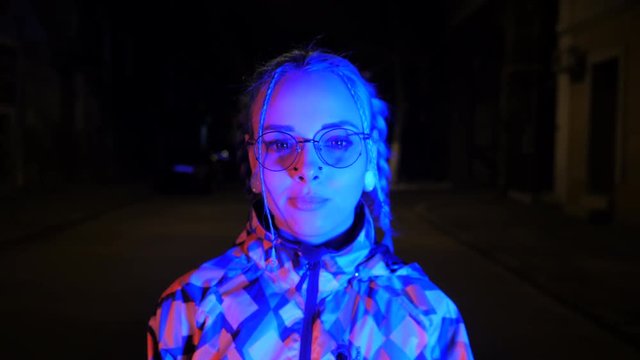 Attractive pretty girl with unusual hairstyle walking on street with light at night. Dyed blue hair in braids. Pensive hipster teenager in glasses chews cud and blows bubbles.