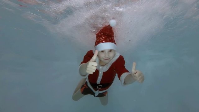 A little boy dressed as Santa Claus swims underwater in the water jets in the pool with his eyes open, smiles, looks at the camera and shows his fingers up. Slow motion. Raw video. 4K. 25 fps.