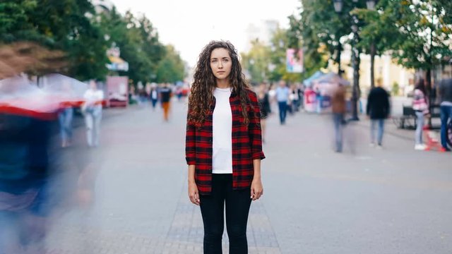 Zoom in time-lapse of beautiful girl with long curly hair standing in street looking at camera when many men and women are walking around in hurry on summer day.