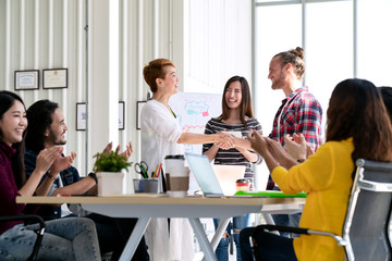 Attractive mature asian woman shaking hand with young beard hipster caucasian with welcome new employee, congratulating successful or happy with business deal while team applauding support together.