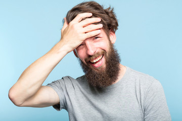 facepalm. happy smiling joyful man covering his face. shame and embarrassment concept. portrait of a young bearded guy on blue background. emotion facial expression. - obrazy, fototapety, plakaty