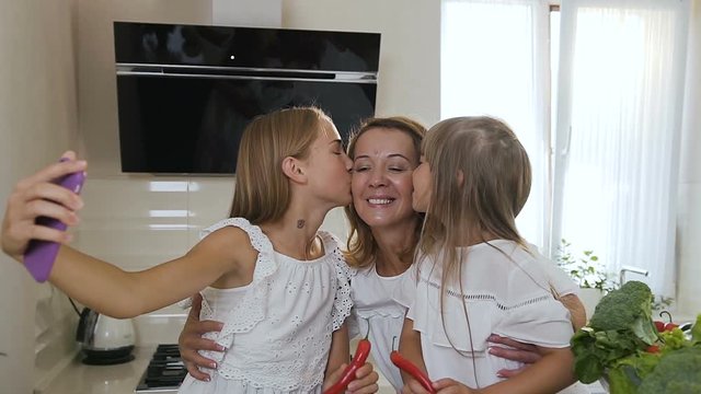 Portrait of a two cute sisters who kiss their beloved and attractive mother in the while cooking eating at home in the kitchen. Girls dressed in white clothes kissing mother and doing selfie photo on