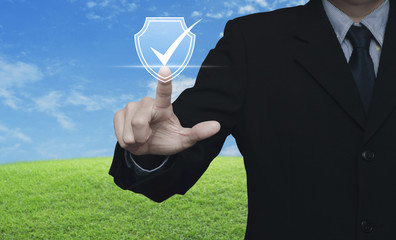 Businessman pressing security shield with check mark icon over green grass field with blue sky, Technology internet cyber security and anti virus concept