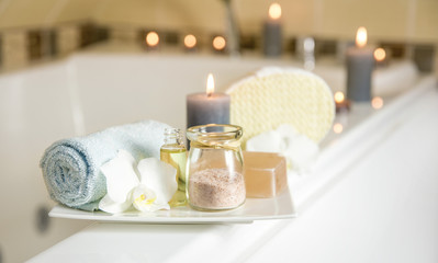 White ceramic tray with home spa supplies in home bathroom for relaxing rituals. Candlelight, salt soap bar, bath salt in jar, massage, bath oil in bottle, blue rolled towel, natural sponge. 