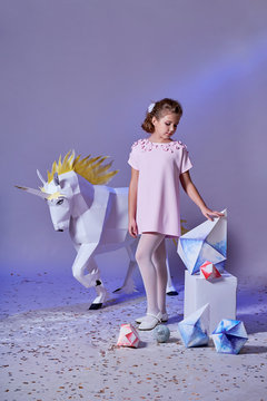 Young kid girl in elegance dress pink. Concept winter, eve, Christmas, new year. Fashion lady teenage poses for children's clothing catalog. White big unicorn origami near. Studio purple background.