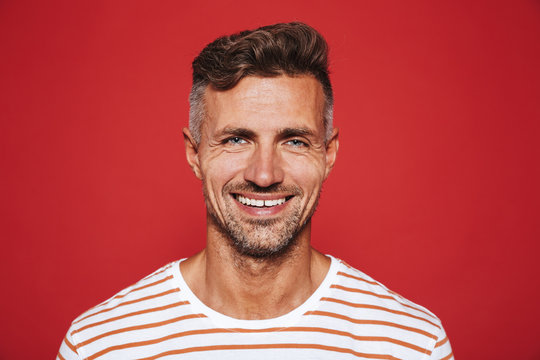 Happy attractive man with stubble in striped t-shirt smiling on camera, isolated over red background