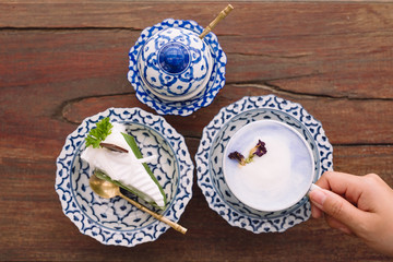 Top view image of a hand holding a cup of hot milk with a piece of cake in an antique Thai traditional benjarong style containers
