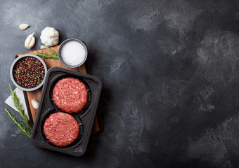 Fototapeta na wymiar Plastic tray with raw minced homemade beef burgers with spices and herbs. Top view and space for text on stone kitchen table background with tomatoes salt and pepper.