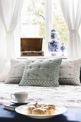 Close-up of a sweet breakfast on a bed in a stylish bedroom interior. Velvet pillows against a bright window draped in white see-through curtains. Real photo.