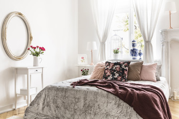 Sweet bedroom interior with a bed covered with a silver throw, a burgundy brown blanket and many cushions. Real photo.