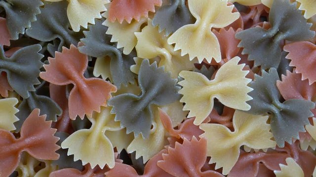 Multicolored pasta background, rotating, top view. Rotation of colored farfalle pasta. Heap of uncooked pasta. Italian food close up. Delicious italian farfalle tricolor pasta, food background