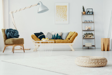 Pouf on rug in white apartment interior with lamp between chair and mustard settee. Real photo