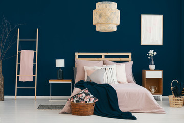 Wicker lamp hanging above double bed in real photo of bedroom interior with dark blue wall, flowers on bedside table and ladder with blanket