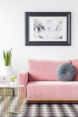 Pillow on pink sofa next to gold table with plant in living room interior with poster. Real photo